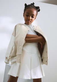 Beige Varsityjacke über einem weißen, gerippten Oberteil und einem plissierten weißen Rock. Der Stoff scheint weich zu sein, mit Knöpfen und Streifen an den Bündchen der Jacke.