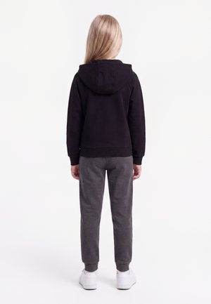 Enfant aux longs cheveux blonds portant un sweat à capuche noir, un pantalon de survêtement gris et des baskets blanches, debout dos à un fond blanc.