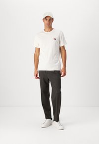 White short-sleeve t-shirt with small logo, paired with dark gray pants and white sneakers. The hat is light tan, fabric texture visible.