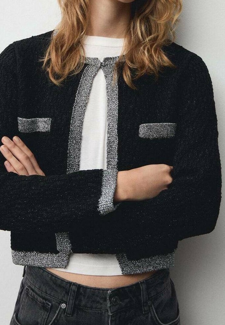 Personne les bras croisés portant un cardigan noir texturé avec une bordure argentée sur une chemise blanche et un jean gris foncé.