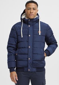 Marineblå puffjakke med hætte, prydet med cremefarvede snore, knaplukning og quiltet design. Den har en blød indvendig foring og lommer.