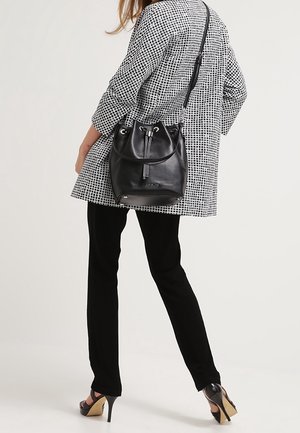 Femme portant un manteau à carreaux noir et blanc et un pantalon noir, portant un sac seau en cuir noir avec une bandoulière.