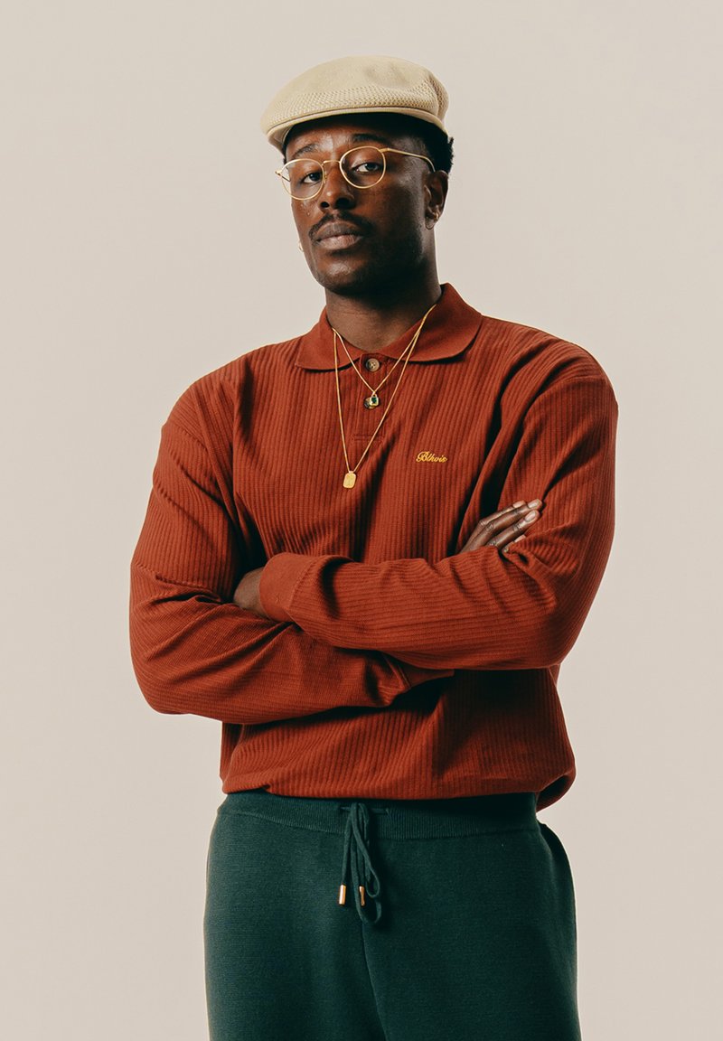 Mężczyzna w beżowej płaskiej czapce, złotych okularach, rudym swetrze, złotych naszyjnikach i ciemnozielonych spodniach, stojący z założonymi rękami.