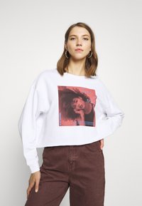 Sweatshirt curto branco com uma estampagem gráfica apresentando uma mulher em tons de vermelho. Mangas longas, decote arredondado e um corte descontraído.