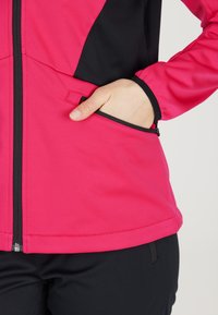 Rosa og sort softshell-jakke med en lynlåslomme, glat tekstur og kontrasterende designelementer på siderne og ærmerne.