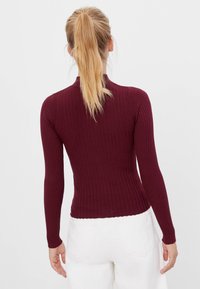 Burgundy-ribbad stickad långärmad topp med hög hals och en kroppsnära silhuett, accentuerad av en scallopkant och texturerad detaljering.