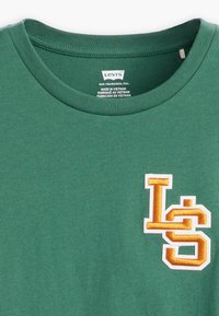 T-shirt en coton vert avec un col rond, arborant un logo "LS" brodé blanc et orange sur le côté gauche. Étiquette visible à l'intérieur du col.