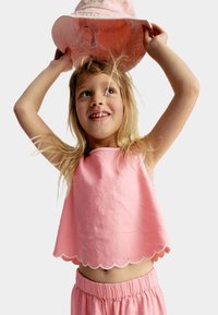 Haut sans manches rose avec un ourlet en forme de vagues, associé à un short avec taille élastique assorti. L'enfant tient un chapeau de seau rose au-dessus de sa tête.