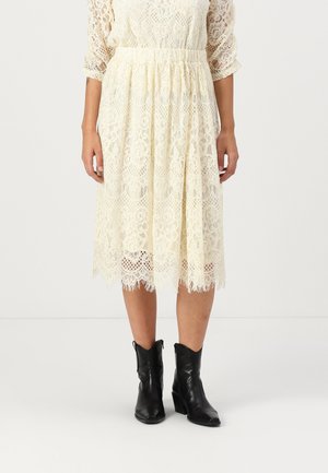 Robe en dentelle crème à manches trois-quarts, taille froncée et ourlet en forme de coquille, assortie à des bottines noires à talon épais.