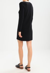 Black long-sleeve dress with a scallop hem, made from a soft fabric. Worn with black sandals, showing a flat heel and ankle strap.