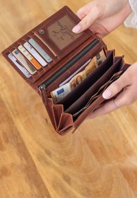 Hände halten eine geöffnete braune Geldbörse mit mehreren Kreditkarten und Euro-Banknoten darin, über einer Holzoberfläche.