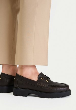 Person trägt beige Hose und dunkelbraune Leder-Slipper mit dicker schwarzer Gummisohle.