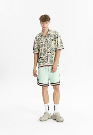 Joven de pie con camisa de manga corta beige con estampado, pantalones cortos a cuadros verde claro con ribete negro, calcetines blancos y zapatillas beige.