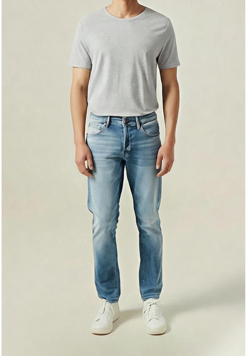 Homme portant un t-shirt gris clair, un jean bleu délavé et des baskets blanches, debout devant un fond neutre uni.