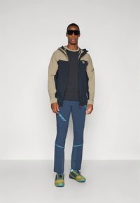 Σετ πεζοπορίας που περιλαμβάνει μπεζ και ναυτικό μπουφάν, μαύρο πουκάμισο, μπλε παντελόνι με λεπτομέρειες, και πράσινα παπούτσια πεζοπορίας. Γυαλιά ηλίου ολοκληρώνουν την εμφάνιση.