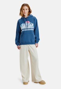 Sudadera azul con un gráfico de montaña, rayas blancas contrastantes, bolsillo frontal, cordones ajustables, combinada con pantalones anchos de color claro.