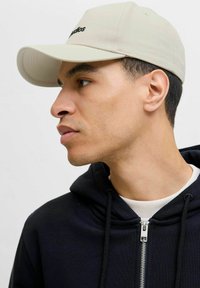 Beige cap with a structured design and curved brim. Features black embroidered logo. Worn with a dark hoodie, showing zipper detail.