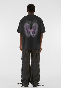 Tricou grafic negru cu un imprimeu mare de fluture purpuriu pe spate, cu detalii estompate, asortat cu pantaloni cargo negri largi.