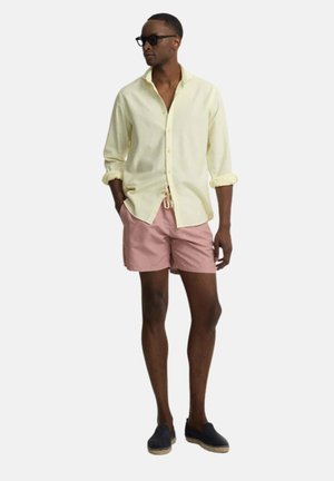 Hombre con camisa de botones amarillo pálido, pantalones cortos rosas, gafas de sol negras y zapatos oscuros sin cordones, de pie con las manos en los bolsillos.