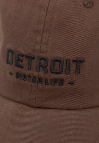 Căciulă maro dintr-un material texturat, cu broderie "DETROIT - MOTOR LIFE" în negru. Cozoroc curbat cu accente de cusătură.