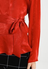 Blusa di raso rosso con maniche lunghe, cintura legata in vita e orlo peplum, abbinata a una gonna nera a quadri. Texture liscia e finitura lucida.