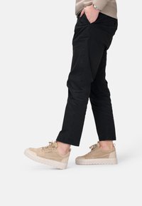 Zapatillas de ante beige con suelas de goma blancas, cerca de pantalones negros. Las zapatillas cuentan con cordones y acentos perforados para mayor transpirabilidad.
