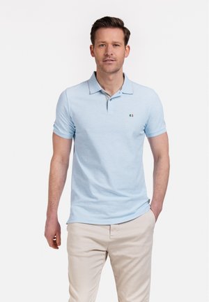 Polo shirt bleu clair à manches courtes, avec un col et des finitions subtiles. Logo brodé sur la poitrine ; associé à un pantalon beige clair.