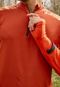 Langärmliges Sportoberteil in einfarbigem Orange mit einem Reißverschlusskragen, ausgestattet mit kontrastierenden, gemusterten Schulterakzenten und Daumenlöchern.