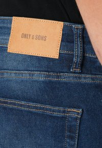 Jeans de mezclilla en azul oscuro con un acabado texturizado. Presenta un parche de cuero marrón en la cinturilla con el logo en relieve "ONLY & SONS".