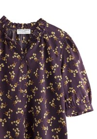 Lilla blomsterbluse lavet af bomuld med en flæsekrave, korte puffærmer og frontknapper. Har et lille mønster af hvide og gule blomster.