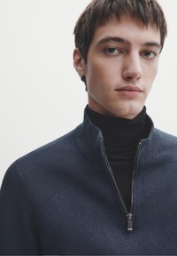 Pull à fermeture éclair bleu marine avec col montant, détails côtelés et fermeture éclair argentée. Superposé à un col roulé noir, texture lisse.