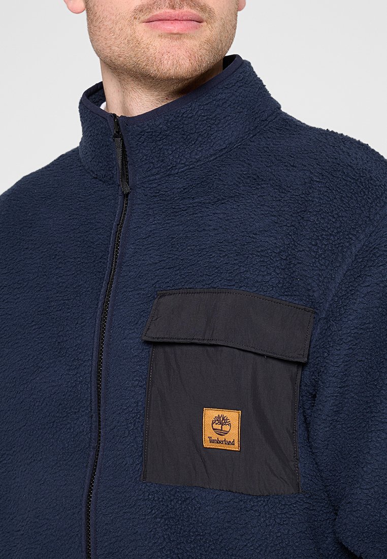Giacca in pile navy con colletto alto e tasca frontale nera; presenta un logo Timberland sulla tasca; materiale testurizzato e caldo.