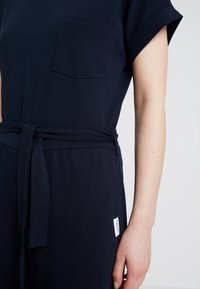 Bezramenný námořnický overal s kapsou na hrudi a páskem v pase, nošený osobou s viditelným pravým ramenem na jednobarevném světlém pozadí.