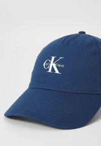 Sötétkék baseball sapka, pamutból készült, ívelt karimával és varrott textúrával, fehér "CK" logóval és "Calvin Klein" felirattal.