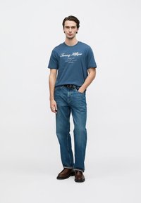 Μπλε βαμβακερό T-shirt με λευκό λογότυπο, συνδυασμένο με τζιν μεσαίου πλυσίματος. Το μοντέλο φορά καφέ παπούτσια και μαύρη ζώνη, στέκεται με αυτοπεποίθηση.