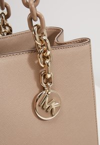 Sac à main beige texturé avec des poignées en chaîne dorée et un pendentif rond en or avec les initiales MK.