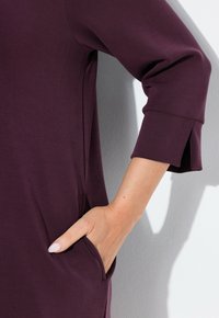 Sukienka z elastycznego materiału w kolorze śliwkowym, z rękawami 3/4, boczną kieszenią i rozcięciem przy mankietach. Gładka faktura, casualowy styl.