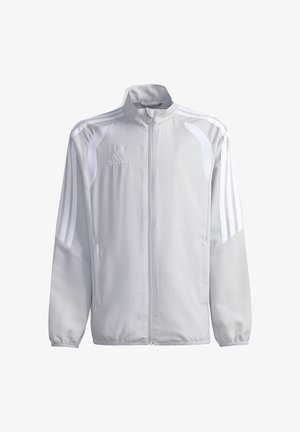 Joven con pelo rizado recogido en un moño alto, que lleva una chaqueta Adidas gris claro con cremallera y rayas blancas en las mangas, de pie y sonriendo.