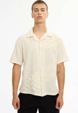 Joven con una camisa beige de manga corta con patrones texturizados de hojas y pantalones negros, de pie sobre un fondo blanco liso.
