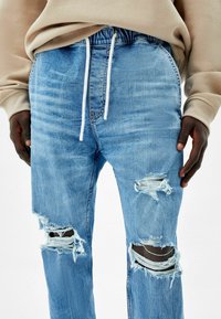 Jeans de mezclilla de un azul claro desgastado con desgarros en las rodillas, que cuentan con una cintura con cordón y un corte relajado. Tejido texturizado con bordes deshilachados.
