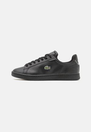 Baskets basses noires avec lacets et semelle noirs, avec un logo crocodile vert et blanc sur le côté.