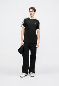 Čierne tričko s bielym lemovaním a tromi bielymi pruhmi na rukávoch, spárované s čiernymi širokými nohavicami a bielymi teniskami. Drží čiernu čapicu.