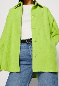 Hellgrüne, übergroße Wollmischjacke mit Kragen, ausgestattet mit großen Knöpfen, getragen über einem weißen Oberteil und kombiniert mit blauen Jeans.
