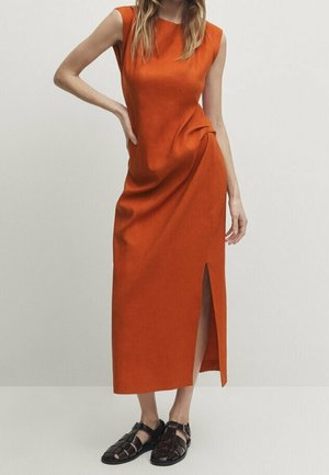Vrouw die een mouwloze oranje midi-jurk met zij-split draagt en zwarte gevlochten gesloten sandalen, staand met één hand op de heup.