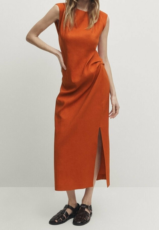 Femme portant une robe midi orange sans manches avec une fente latérale et des sandales noires tissées à bout fermé, debout avec une main sur la hanche.