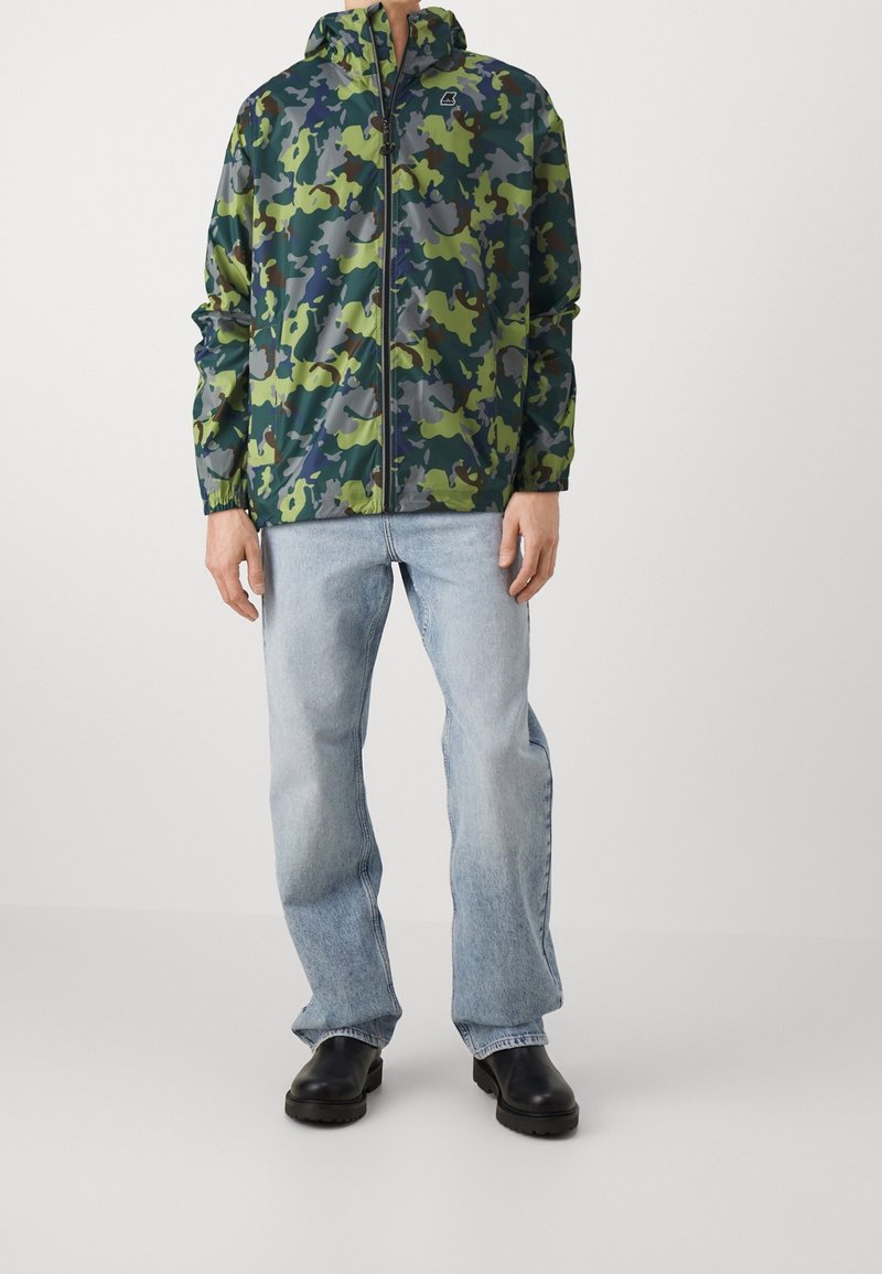 Homme portant une veste zippée camouflage verte et grise, un jean droit bleu clair et des bottes noires sur un fond uni.
