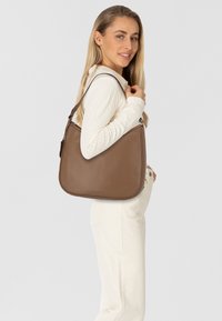 Sac à bandoulière en cuir marron avec une texture lisse, une forme courbée, une sangle ajustable et un logo discret en bas.