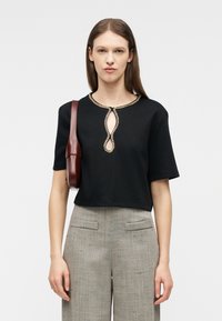 Mujer con una blusa negra de manga corta con escote en forma de ojo de cerradura adornado con joyas, pantalones grises de cintura alta y un bolso marrón de hombro.