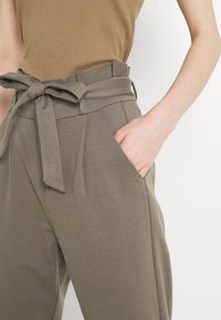 High-waisted, olive green trousers made of soft fabric with a tied belt detail, pleats, and side pockets.