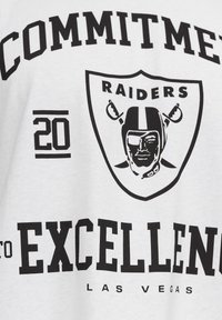 Camiseta blanca con texto impreso en negro que dice "COMPROMISO con la EXCELENCIA" junto con "RAIDERS" y un logo que representa un casco de fútbol americano.